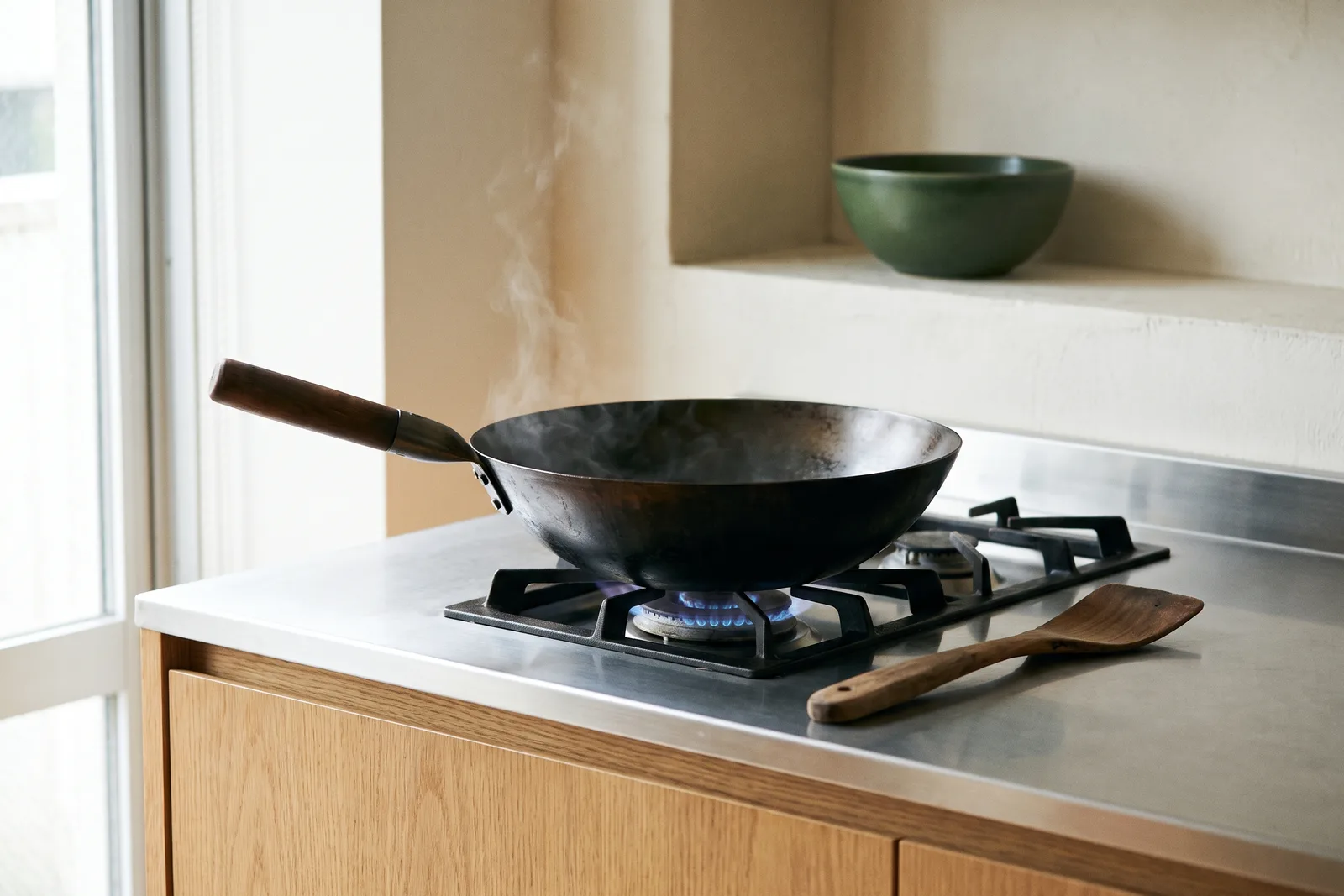 A seasoned carbon steel wok steaming over a blue gas flame on a stainless and oak kitchen counter.